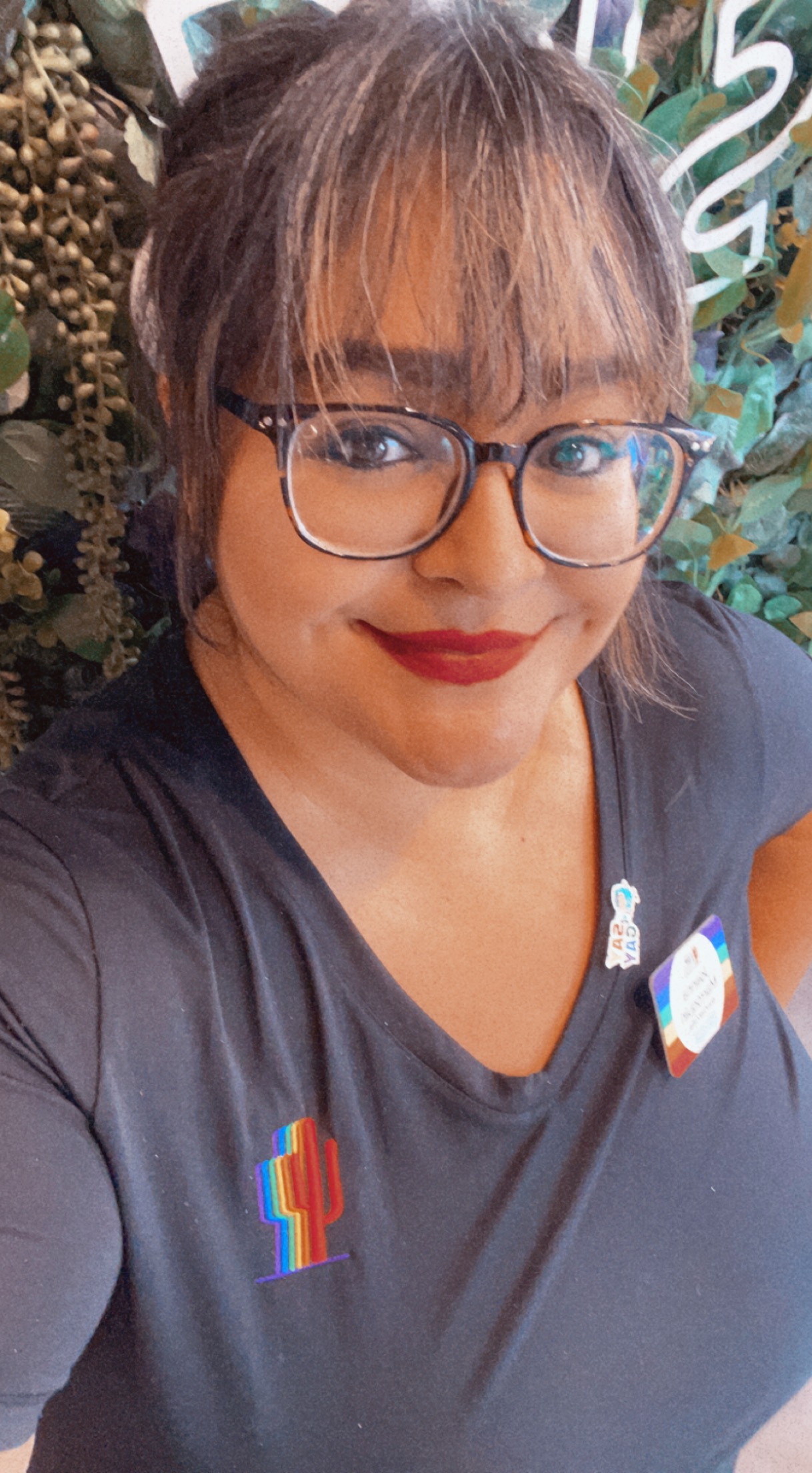 Brown skinned woman with red lipstick, classes, and bangs looks up at the camera smiling wearing a Gaymber t-shirt. 