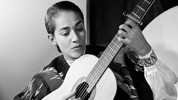 Black and white image of a woman with dark hair pulled back looking at an acoustic guitar as she plays it