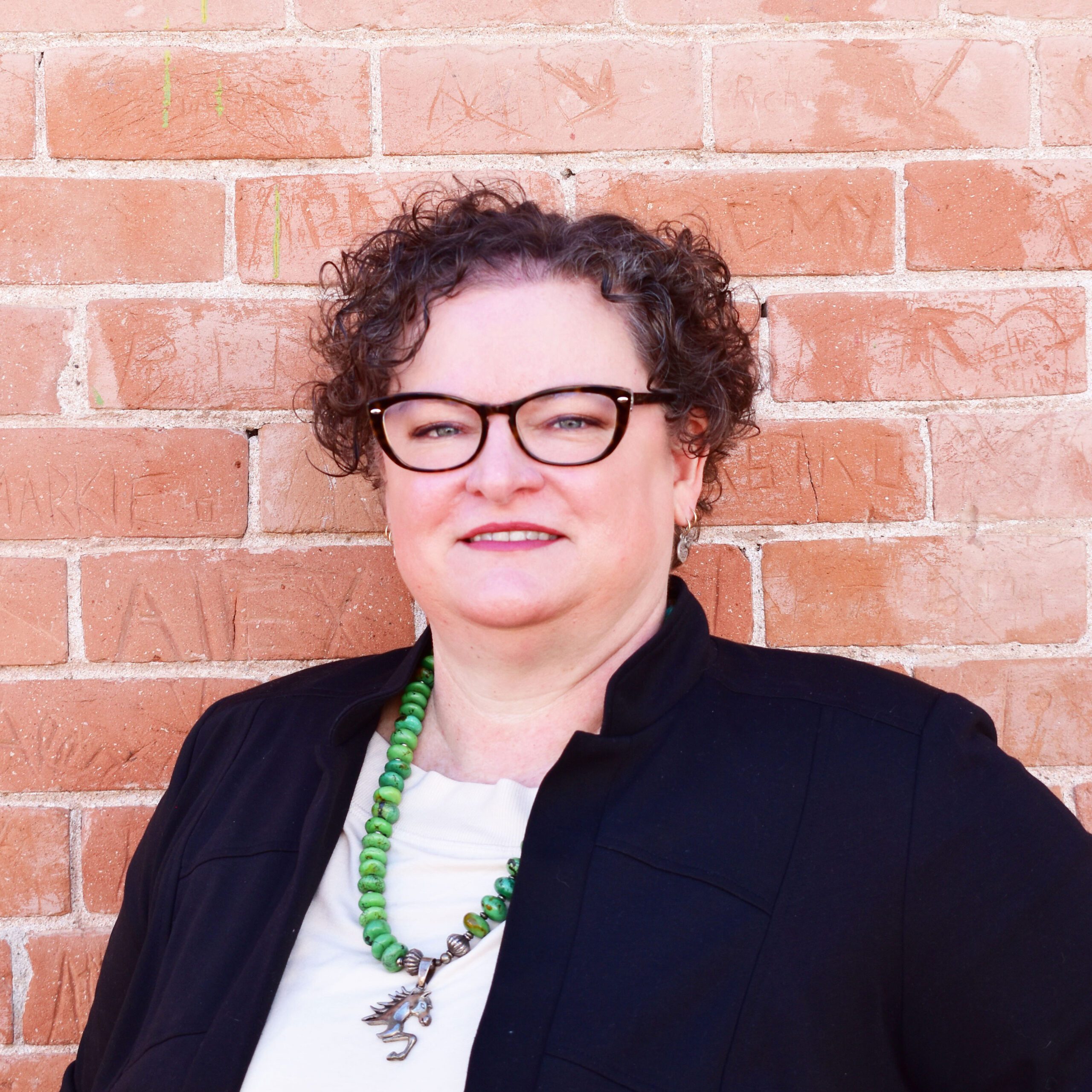 Lisa Bowers, Gaymber President, a light-skinned woman with curly hair and glasses stands in front of a brick wall